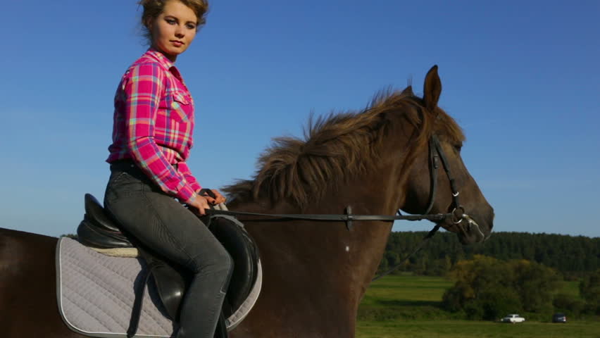 Attractive young girl riding a horse. The horse is standing still, the Mare turns her head looking at the camera. Medium shot, side view, summer, dry season, outdoor, natural light, real time