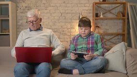 The difference of generations. Elderly man with glasses sitting on sofa working on laptop, young boy playing on tablet. grandfather and grandson. Home comfort, family idyll, cosiness concept 60