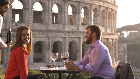 Elegant waiter serving pouring glass of red wine to romantic couple sitting at restaurant table in front of colosseum in rome at sunset