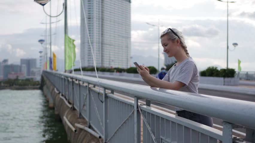 Slim woman with a pigtail blonde in a gray T-shirt standing on the bridge and using smartphone over background sea and city. Girl touching screen