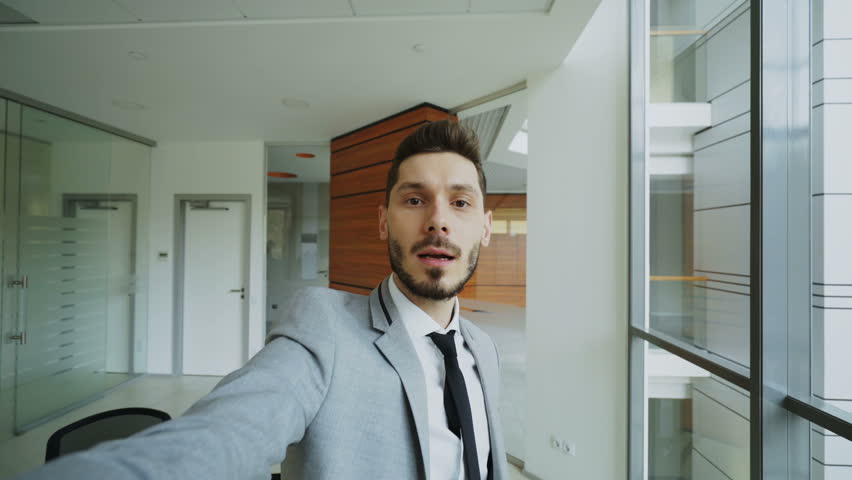 POV of young businessman in suit having online video chat using smartphone camera and talking to his colleagues in modern office
