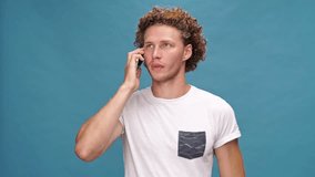 Smiling curly man in t-shirt talking by smartphone over blue background