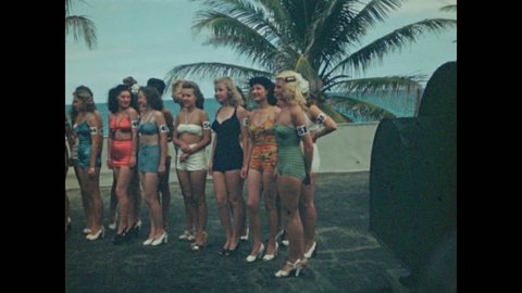 1940s: Beauty pageant, long line of women with numbered armbands stand in swimsuits in front of palm trees, pose. Women all begin to walk forward. Swimmers do the butterfly.