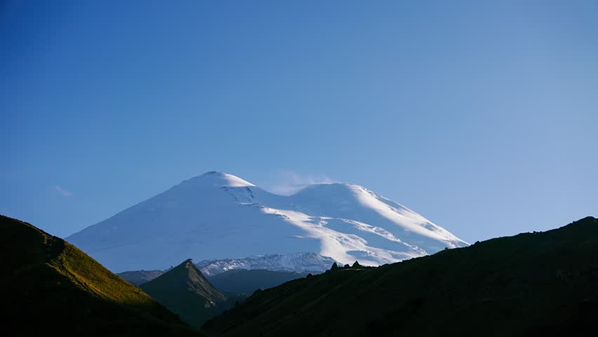 Mount Elbrus in the light of the setting sun. The twin summits of this dormant stratovolcano dominate the frame, their snow-covered volcanic domes catching the last warm rays of daylight while the surrounding Caucasus slopes fall into deepening shadow. The western peak, the highest point in Europe at 5,642 meters, stands slightly taller than its eastern counterpart, with both cones separated by a glacial saddle visible in the composition. The sky transitions from deep blue to soft twilight purple as scattered clouds drift past the upper ridges, and the vast glacial fields that feed more than twenty rivers shimmer in the fading light. The scene captures the mountain