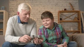 The grandson teaches grandfather to play the console game. An elderly man is learning how to play a fighting video game. Home comfort, family idyll, cosiness concept, difference of generations 60 fps