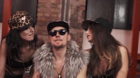 Wealthy caucasian rapper man in fur vest with glasses and snapback with sexy girls in underwear sitting on bed covered in money rapping