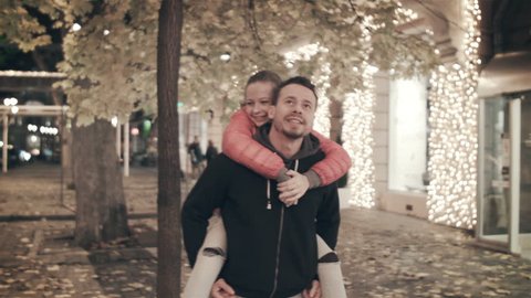 A happy family walks the evening in the city on holidays. The father carries on his back a daughter who tears a leaf from an autumn tree