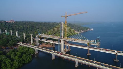 Construction of a huge cable-stayed bridge over the river. Cars go on the old part of the bridge. Tower cranes over the bridge across the Mandovi river in Goa, India. Shooting with a drone.