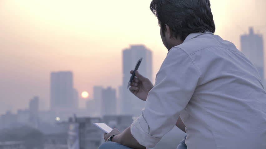 A young man holding a notepad and pen  looking at a rising sun and city skyline 