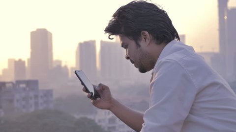 A young man typing a text message on a mobile phone against the vibrant and beautiful city skyline. A young attractive male using smartphone or cellphone to type a SMS or email against setting sun  