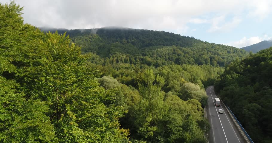 Aerial view from flying copter over beautiful sunny mountains covered with trees near the road full of driving cars.