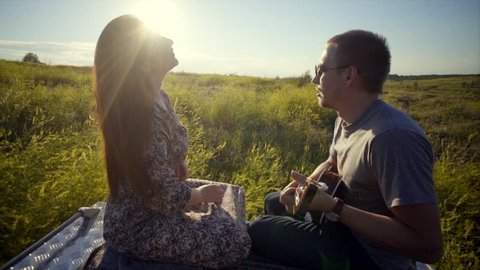 Young man and woman siting on the back of pickup track in field. Couple in sunset light man playing on ukulele woman hearing music and singing