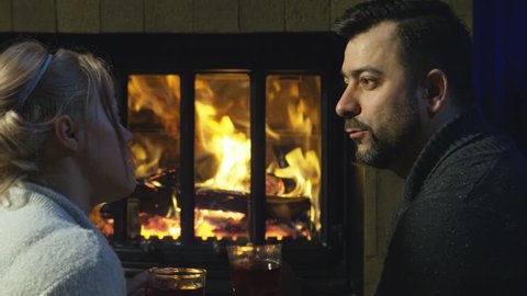 Romantic couple relaxing on the floor in front of a burning log fire in winter with a view from behind of their feet in slippers as they chat and sip coffee or gluhwein with a Christmas gift between.