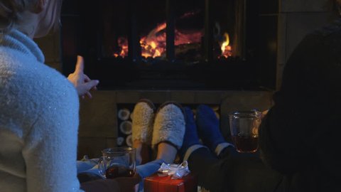 Romantic couple relaxing on the floor in front of a burning log fire in winter with a view from behind of their feet in slippers as they chat and sip coffee or gluhwein with a Christmas gift between.