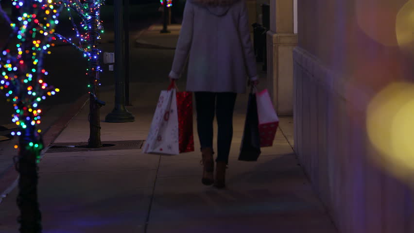 Woman walking down sidewalk with shopping bags along trees with Christmas lights on.