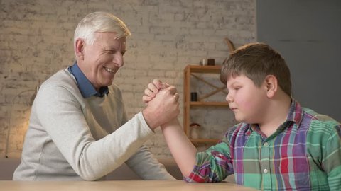Grandfather and grandson arm wrestle in a cozy room at home. Measuring forces in arm wrestling. Young fat child won. Home comfort, family idyll, cosiness concept, difference of generations, close up