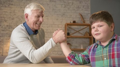 Grandfather and grandson arm wrestle in a cozy room at home. Measuring forces in arm wrestling. Old man won. Home comfort, family idyll, cosiness concept, difference of generations, close up. 60 fps