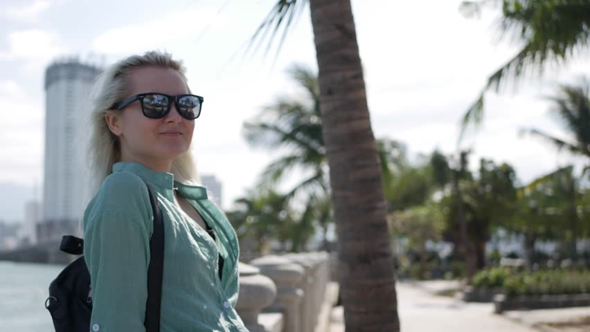 Happy caucasian woman with long blonde hair in sunglasses and green shirt standing and smiling near palm tree on a park and sea background. Travel concept