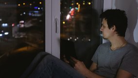 Man sitting on the windowsill in the dark night and looking at street. Using tablet for web.