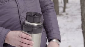 Open the thermos. The black cover is unscrewed by hand. The iron hull. On the background of the winter forest. Body part. The jacket is blue. The light of the day. Big shot. 4K video