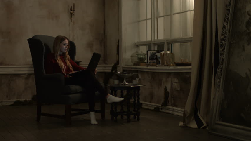 Adorable young woman streaming video content on line using laptop pc and gently smiling while sitting in vintage armchair in retro interior in the evening. Cheerful girl working with laptop at night.