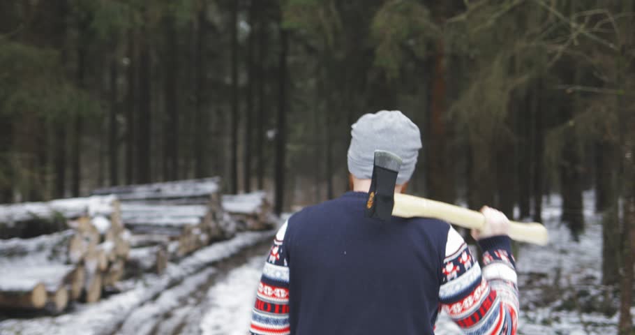back view, strong man / lumberjack holding ax (axe, hatchet) in his hands and coming to the winter forest on the snowy road