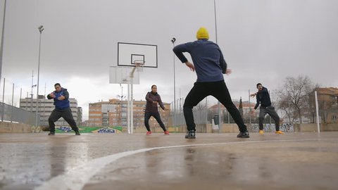 Street workout training. Sport group of young multi ethnic people practicing tai chi or qigong on the outdoor basketball court in the rain. The coach with yellow knit cap explains proper movements