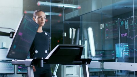 Beautiful Woman Athlete with Electrodes Connected to Her Body Walks on a Treadmill in a Sports Science Laboratory. In the Background High-Tech Laboratory with Monitors Showing EKG. RED EPIC-W 8K