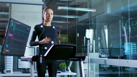 In Scientific Sports Laboratory Beautiful Woman Athlete Runs on a Treadmill with Electrodes Attached to Her Body, Monitors Show EKG Data on Display. Slow Motion. Shot on RED EPIC-W 8K Helium  Camera.