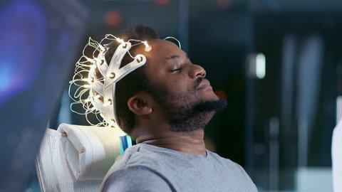 In Laboratory Man Wearing Brainwave Scanning Headset Sits in a Chair with Closed Eyes. Monitors Show EEG Reading and Graphical Brain Model. In the Modern Brain Study Neurological Research Laboratory.