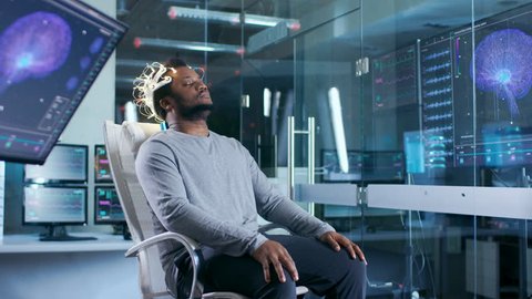 In Laboratory Man Wearing Brainwave Scanning Headset Sits in a Chair with Closed Eyes. In the Modern Brain Study Neurological Research Laboratory. Monitors Show EEG Reading and Graphical Brain Model. Stock Video