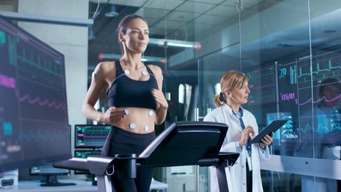 Woman Athlete Runs on a Treadmill with Electrodes Attached to Her Body, Female Physician Uses Tablet Computer and Controls EKG Data Showing on Laboratory Monitors. Shot on RED EPIC-W 8K Camera.