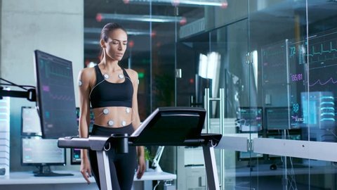 Beautiful Woman Athlete Wearing Sports Bra with Electrodes Connected to Her, Walks on a Treadmill in a Sports Science Laboratory. In the Background Laboratory with Monitors Showing EKG data. 4K UHD.