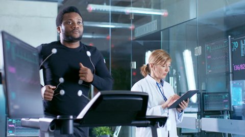 Male Athlete Runs on a Treadmill with Electrodes Attached to His Body while Sport Scientist Holds Tablet and Supervises EKG Status. Laboratory with High-Tech Equipment. Shot on RED EPIC-W 8K Camera.