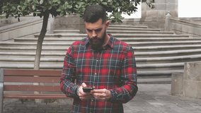 Modern hipster with red checkers flannel shirt recording video with mobile on the street. Young full bearded man taking photos with cellular in old town Vegueta, Las Palmas. City tour visit concept