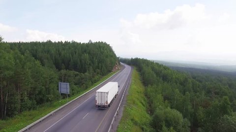 One white truck driving along the freeway amidst a dense forest in the mountains. Summer, sunny, beautiful sky with clouds. Aerial drone view.
