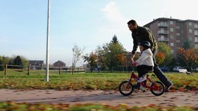 father teaching daughter to ride bike at urban park. Child girl learning biking with the dad's help.Family and childhood concept.Sunny autumn day.Side gimbal follow 4k video 