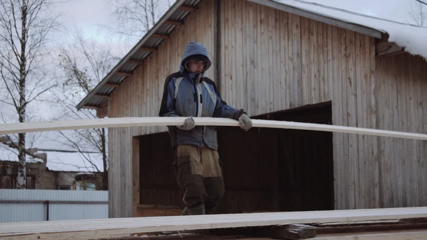 Caucasian man porter in dirty clothes carrying wood plank at sawmill factory yard on cold winter day
