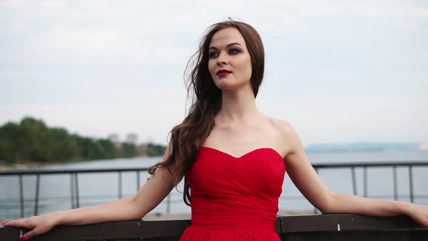 Pretty young woman with red lips is leaning back on handrails of embankment. Sea water and coast with green trees are in background, girl is looking and having a little smile