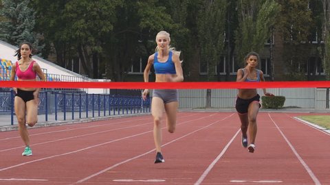 Female marathon runners crossing finishing line on professional sports arena. Fitness women racing competition at stadium, victory celebration, achievement. Motivated blond girl winning speed contest
