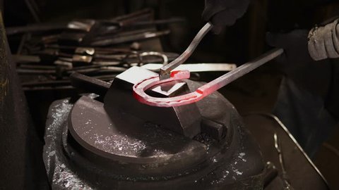 Worker is holding heated blank of horseshoe on a anvil of hydraulic hammer in a workshop of forge. Close-up of product, hands of man and instruments