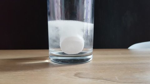 Aspirin or effervescent pill dropping into a glass of water on the table in dark background, Pill drops into the water, tablet dissolves in water with fizzy.
