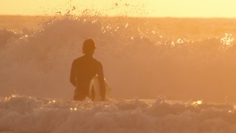 SLOW MOTION: Large ocean waves violently crash and break in front of golden lit surfer. Unrecognizable male surfer walks towards ocean waves with surfboard in his hand on a beautiful golden evening.
