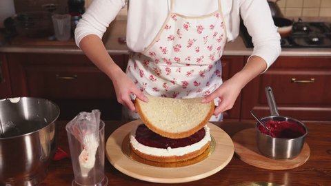 Confectioner woman is assembling layer cake. She is laying over whipped cream layer a baked cake, in home kitchen
