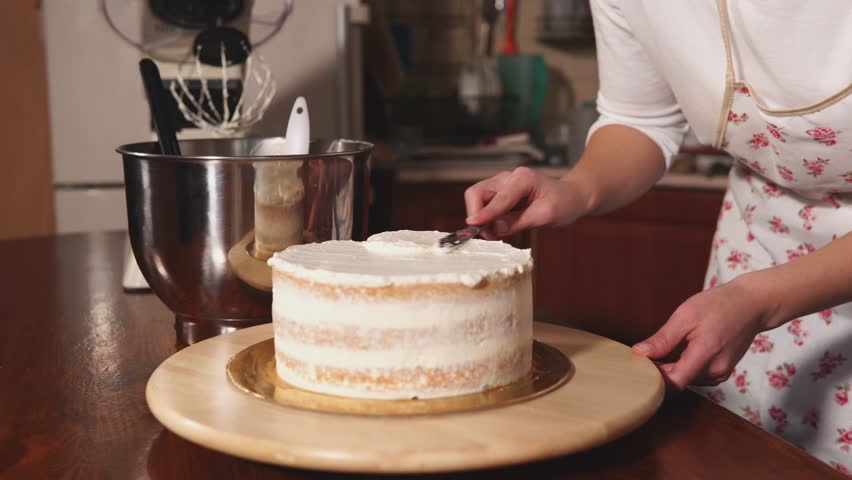 Pastry chef is applying creamy cream on an upper layer of biscuit cake. Talented woman is making authors cakes in her own kitchen