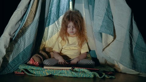 A little girl in pajamas is sitting and reading a book to her toy hare in a wigwam tent in a game room in the evening before going to bed.
