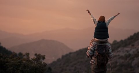 Successful father hiker standing on mountain peak looking at view with son on shoulders