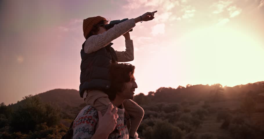 Son on hiker father's shoulders using binoculars and looking at nature on mountains at sunset