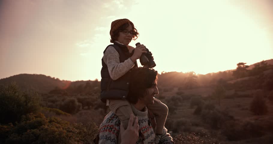 Young son on hiker father's shoulders using binoculars and looking at mountain view at sunset