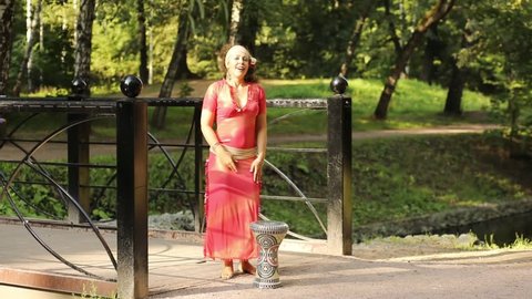 Beautiful woman in red dress dance bellydance near darbuka on bridge in park at sunny day.